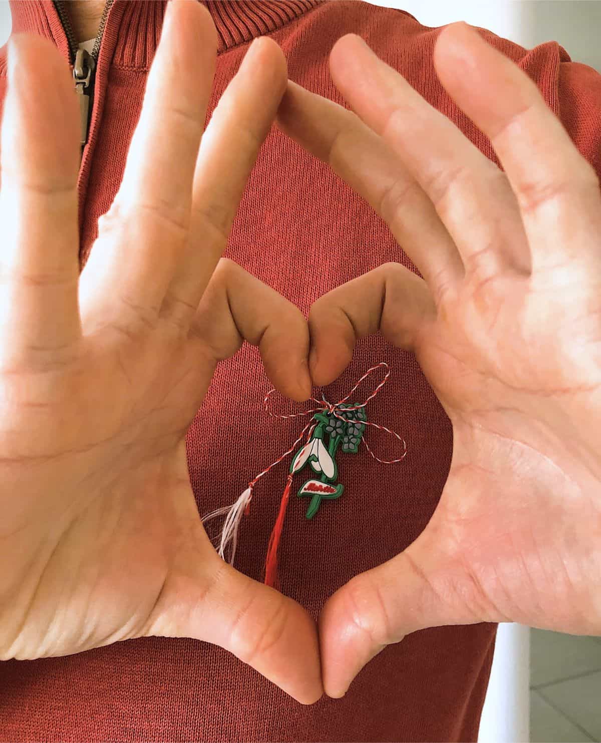 Person holding hands in heart shape around a red and white Martisor charm pinned to a sweater.
