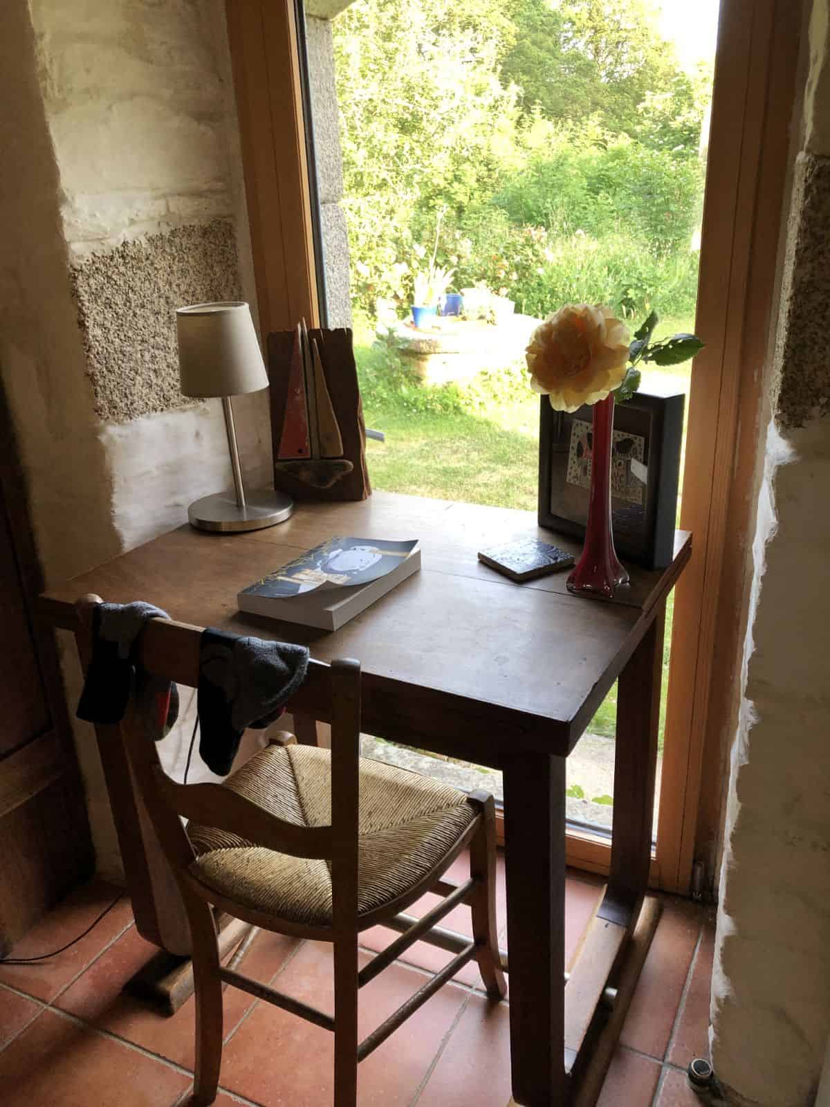 Wooden desk with book, chair, and flower by a window overlooking a garden in daylight.